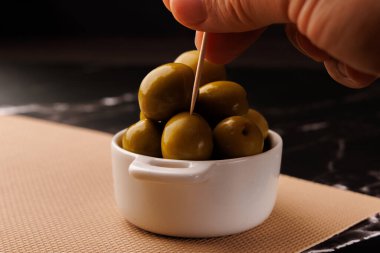 Bir insan siyah mermer yüzey ve siyah arkaplan üzerinde bir tabağın yanında bir sopayla bir zeytin ve yağ şişesi seçer.