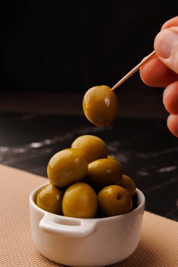 Bir el, siyah mermer bir masanın üzerindeki yağ şişesinin yanında yumuşak siyah bir arkaplan ile zeytin tabakasıyla etkileşir.