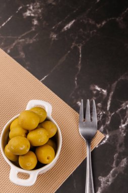 Yukarıdan: bir kasede zeytin, bir yağ şişesi ve siyah mermer bir masanın üstünde tahta sopa kullanan bir el.