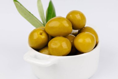 Yeşil zeytinler beyaz bir seramik kasenin içinde, parlak beyaz bir yüzeye vurulan zeytin dalının yanında..
