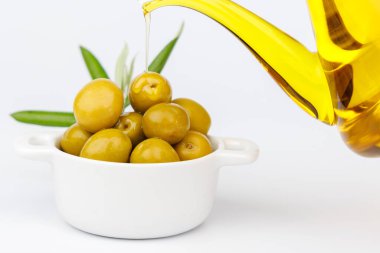 Taze yeşil zeytinyağı, beyaz yüzeydeki yeşil zeytin dalının yanında cam bir şeker kutusundan dökülüyor.