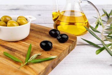 Olgun zeytinlerin yanında temiz bir şişe zeytin yağı ve siyah ahşap üzerinde zeytin dalları, temiz beyaz bir arka plan ile.