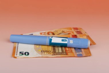 Semaglutide kalemleri, yüksek uyuşturucu fiyatları ve sağlık hizmetlerinin mali ağırlığını öne süren, kırmızı bir arkaplan ile nakit para banknotları üzerinde dinleniyor.