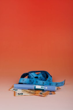 Nakit para ve mezura üzerine konmuş sendromlu dolma kalemler, kırmızı tonlu bir ayarda tıbbi kilo kaybının artan maliyetlerini yansıtıyor.