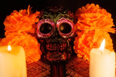 Metalik çiçeklerle süslenmiş siyah bir kafatası, manolyalar ve çok renkli mumlarla dolu canlı bir sunağın üzerinde duruyor..