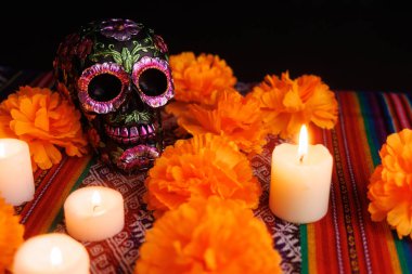 Metalik çiçeklerle süslenmiş siyah bir kafatası Dia de los Muertos için folklorik sunakta beyaz mumların ve manolya çiçeklerinin arasında duruyor..