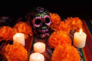 Metal çiçekli siyah bir kafatası sunağın üzerinde duruyor. Beyaz mumlar ve manolya çiçekleri Dia de Muertos anısına..
