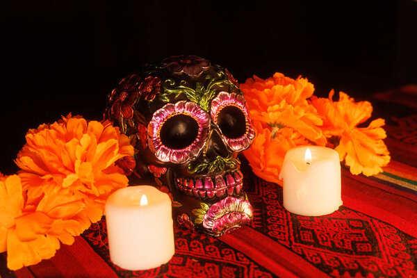 Мексиканский алтарь с черным черепом, раскрашенным цветочными узорами, белыми свечами и магнолиями поверх традиционной ткани.