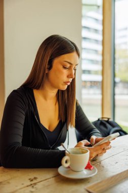 Güzel genç bir kadın, eski bir kafede kahve içerken ahşap bir masada cep telefonunu kullanıyor.