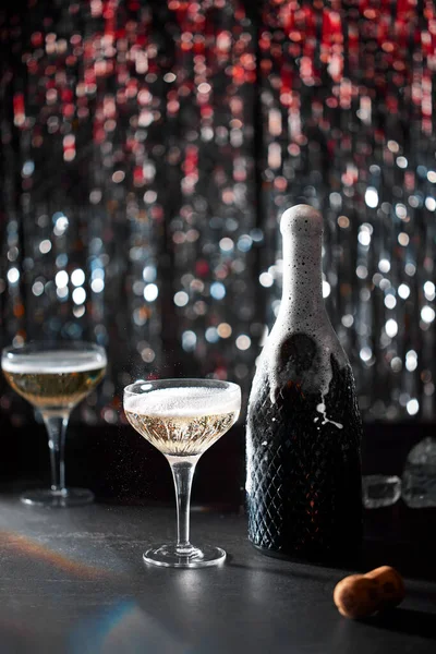 Image of celebration. The champaign bottle splashing next to which are two glasses of champaign. The background of bokeh with colors red and silver. Dark grey stone table.