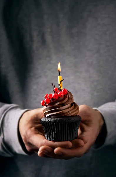Çikolata kremalı kek tutan bir erkek gövdesi. Doğum günü mumu yanıyor. Kırsal süslenmiş kırmızı böğürtlenli kek ve biraz çikolata dalı. Adamın gri kazağı var..