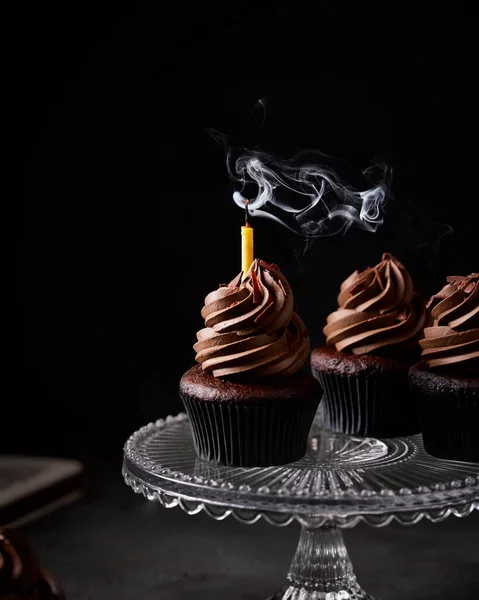 Three chocolate cupcakes on a glass cake stand. There is candle in one cupcake that has been blown out and there is still candle smoke visible.
