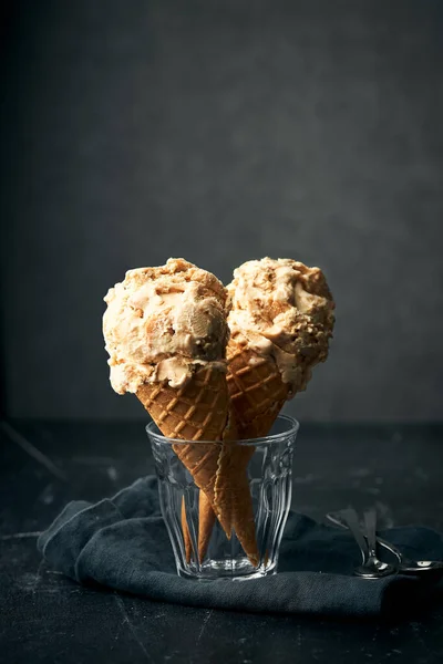 Waffle külahında iki deniz tuzu karamel dondurma pamuk peçetenin üzerinde bir bardakta duruyor. Gri arkaplan ve siyah taş masa üstü. Yan açıdan dikey yaşam tarzı görüntüsü. Kopyalamak için boşluk.