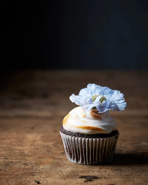 Üzerinde büyük mavi bir çiçek olan kremalı bir çikolatalı kek. Köy dokusundan yapılmış eski ahşap masa. Siyah arka plan. Kopya için alanı olan karamsar dikey natürmort resim.
