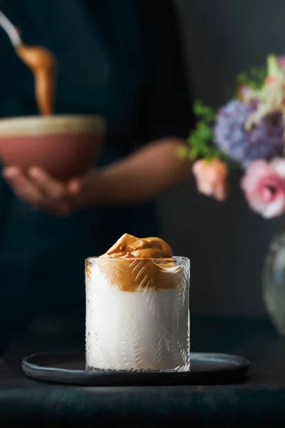 Siyah seramik tabakta Dalgona kahvesi. Arka planda dalgona köpüğünü geleneksel seramik bir kasede hazırlayan bir kadının bulanık gövdesini görebilirsiniz. Masanın üzerinde çiçekler var. Dikey görüntü. Koyu arkaplan. 