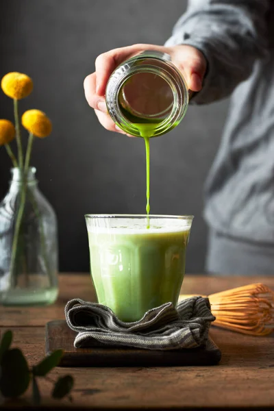 Bir bardak süte kibrit çöpü dökerken kesilmiş. Matcha latte hazırlığı. Ahşap masa ve gri arka plan. Oturma odası mı, yemek odası mı?.