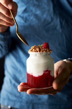 Ahududu reçelli sağlıklı yoğurt ve bir avuç içinde duran tahıl gevreği. Başka bir el kaşıkla kavanoza doğru uzanıyor. İnsan elementi ile hareket halinde dikey görüntü. 