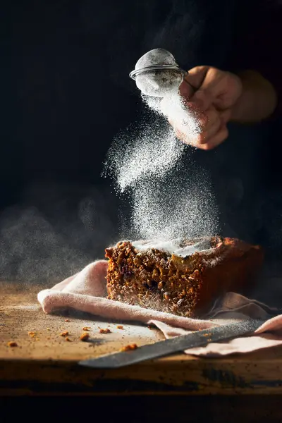Köy ahşap bir masada dilimlenmiş muz ekmeği ve siyah arka plan. Kırpılmış el üstüne şeker tozu sürüyor. Dikey natürmort resim. Kopyalamak için boşluk.