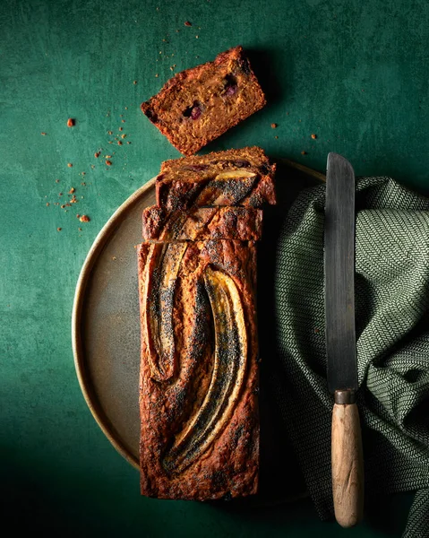 Koyu yeşil desenli arka planda dilimlenmiş muzlu ekmeğin üst görünümü. Kenarında bıçak ve yeşil bir peçete var. Dikey natürmort.