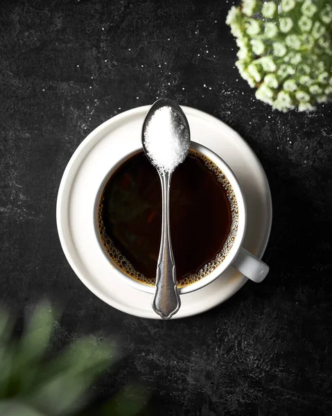 Siyah desenli arka planda, beyaz bir fincanda siyah bir kahvenin üst görüntüsü. Fincanın üstüne bir kaşık şeker. Görüntüde derinlik yaratmak için ön planda bulanık çiçekler var. Kopyalamak için boşluk.