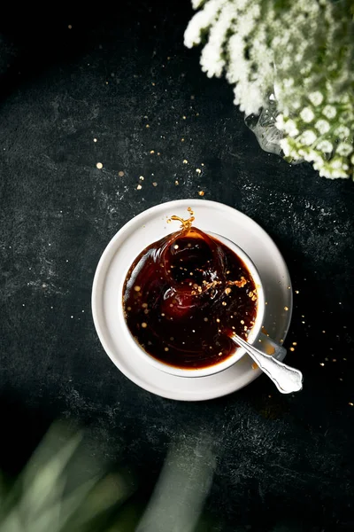 Kahve fincanındaki sıçramanın dikey görüntüsü. Beyaz fincanda sade kahve. Siyah desenli arka plan. Görüntünün derinliğini oluşturmak için köşedeki bulanık çiçekler. 