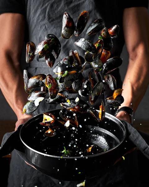 Midyelerin hazırlık görüntüsü. Erkek elleri, onları karıştırmak için siyah tencereden midyeleri havaya fırlatıyor. Etrafta damlalar var. Koyu siyah arkaplan. 