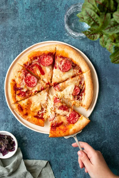 Kırpılmış el, bir dilim pizzayı kek sunucusuyla çekiyor. Peynir esneme hareketi. Mavi desenli arka plan. Etrafta bir bardak su ve çiçek gibi aksesuarlar. Üst görünüm yassı yaşam tarzı resmi.