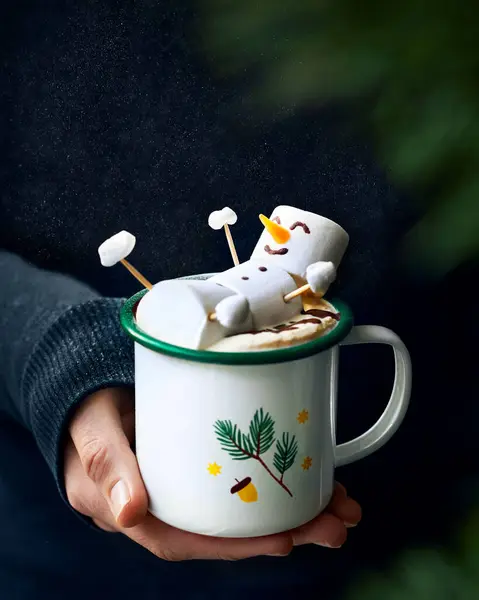 Sıcak kakaonun üzerinde kış temalı bir kupada yenebilir şekerleme kardan adam yatıyor. Bir kadının eli tutuyor. Siyah siyah arka plan. Sıcak kış dikey hayat fotoğrafı. 