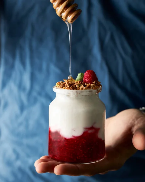 Bal, elinde tutulan yoğurt parfünün üstüne bal damlatılıyor. Arka planda mavi gömlekli erkek gövdesi. Hareketli koyu dikey yemek fotoğrafı. 