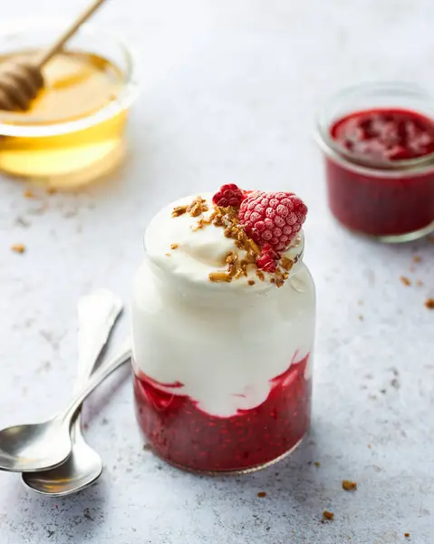 Granola ve ahududu reçelli yoğurt parfe. Beyaz taş arka planda granola kırıntıları, kaşıklar, kavanozda bal ve arka planda ahududu reçeli. Dikey yaşam biçimi.