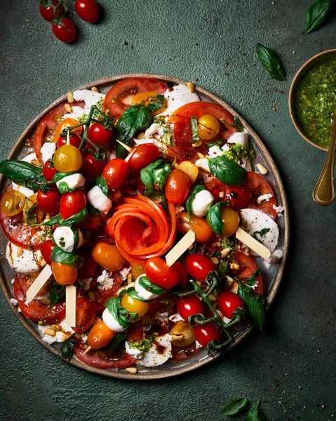 Fesleğen, mozzarella ve çeşitli boyutlarda domateslerden oluşan büyük bir tabak caprese salatası. Koyu yeşil arka plan. Salatanın yanında pesto. Şişler. Dağınık arkaplan, yaşam tarzı resmi.