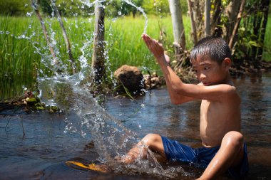 Asyalı genç çocuk yeşil pirinç tarlalarının ve gündoğumlarının arka planında nehirde su oynayan sevimli ve büyüleyici çocuk, su oynayan çocuk, thai çocuk yaşam tarzı, mutlu, eğlenceli, neşe.