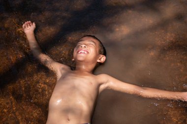 Asyalı genç çocuk gülümseyerek tatlı ve büyüleyici bir şekilde suda oynar ve eğlenir. Genç Asyalı çocuk arka plandaki suyun yüzeyinde yüzer ve tatlı bir güneş ışığı hisseder..