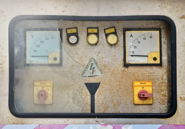 A damaged and rusted electrical control panel has broken glass shards against a soft sunlight background.