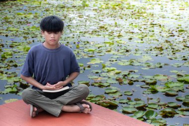 Asyalı çocuk nilüfer havuzunda ve gökyüzü güneşinin altında meditasyon yapıyor, yakın plan çocuk oturuyor, mavi çocuk oturuyor, çocuklar rahatlıyor, konsept meditasyon ve rahatlama, bahçe Thai güzel arka plan, nehir kenarı.