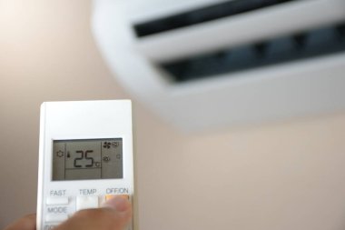 Beyaz odadaki klimayı 25 derecede açmak için uzaktan kumandayı tutarken, arka plandaki elektrik ve elektrik cihazlarını korumak için yumuşak güneş ışığı kavramı bulanıklaştı..