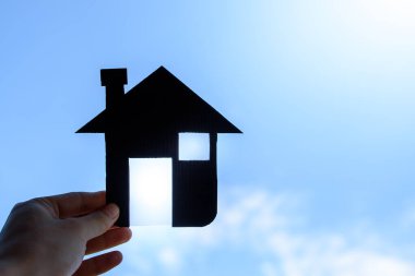 El ele tutuşan küçük siyah model bir ev. Parlak ışık alan bir ev. Mavi gökyüzünde emlak ve sigorta için arka planı bulanık..