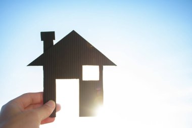 Mavi gökyüzüne karşı minyatür siyah bir ev ya da ev tutan bir adam ve yumuşak güneş ışığı. Emlak ve kiralık ev için güzel bir arka plan..