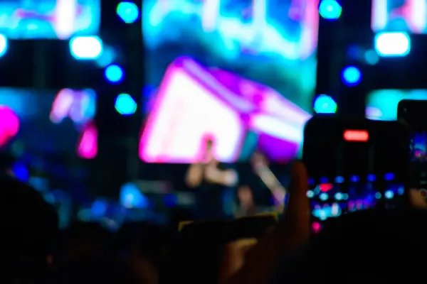 Gece konserinde renkli spot ışıklarıyla video kaydeden akıllı telefonu tutan bir elin bulanık görüntüsü. Geceleri sahne ışıkları altında konser sahnesi.