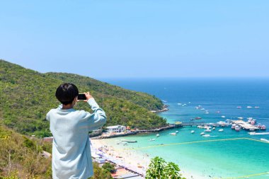 Akıllı telefon tutan adam, tropik bir sahil ve dağların tepesindeki bir adanın fotoğrafını çekiyor. Koh Larn Adaları, Tayland 'da beyaz kumlu sahil manzaralı turkuaz su.