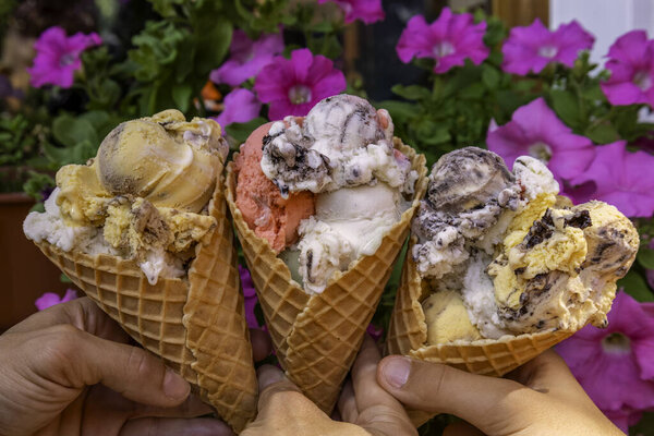 Три разноцветных совок мороженого в вафельных рожках, держащихся за руки. Натуральное красочное мороженое, держащееся в руках. Мороженое в шашлыках, идеальный десерт, закуска. Мороженое на фоне цветов.