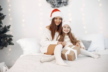 İnsanlar Noel için hazırlanıyor. Yatakta kızıyla oturan bir anne. Aile Noel Baba şapkası takıyor. Beyaz elbiseli küçük kız.