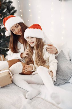 İnsanlar Noel için hazırlanıyor. Yatakta kızıyla oturan bir anne. Aile Noel Baba şapkası takıyor. Beyaz elbiseli küçük kız.