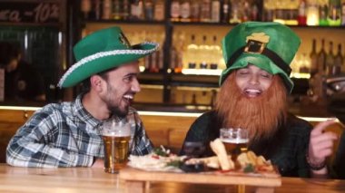 The company of young people celebrate St. Patricks Day. They have fun at the bar. They are dressed in carnival headgear.