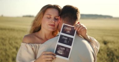 Pregnant woman with husband holding ultrasound baby image. Loving man hugging his pregnant wife from behind in the field. The concept of pregnancy.