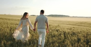 A pregnant woman in a beautiful long dress walks through the field with her husband in a wheat field at sunset. Future parents.