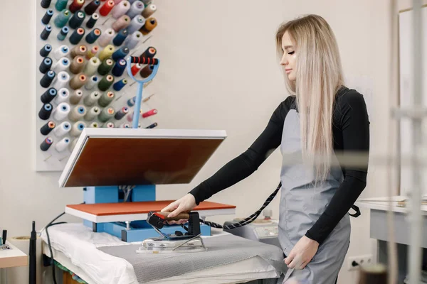 Young female dressmaker standing in tailoring studio near the wall with spools of thread. Blonde woman wearing grey apron. Woman standing near special ironing machine.