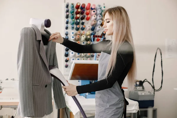 stock image Young female seamstress standing in tailoring studio and ironing a jacket on a dummy. Blonde woman wearing grey apron. Woman working in atelier.