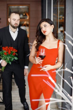 Romantik bir akşam yemeğinde kırmızı elbiseli çekici genç kadına odaklan. Bir buket kırmızı gülle arkada duran adam. Güzel bir çift modern restoranda birlikte vakit geçiriyor..