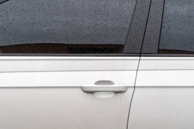 Yağmurlu Gün Sürücüsü: Sağanak Altında Beyaz Araba 'nın Kapanışı. Sağanak yağış altında beyaz bir araba. Pencere ve kapılara yapışmış yağmur damlaları, parlak beyaz yüzeye karşı büyüleyici bir desen oluşturuyor.